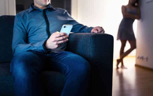 Man seated looking at his phone with a woman in the background appearing suspicious.